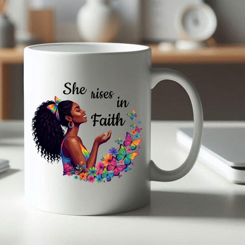 May include: White ceramic mug featuring a colorful illustration of a woman with curly black hair, butterflies, and flowers. The mug has the text "She rises in Faith". The handle is on the right side.