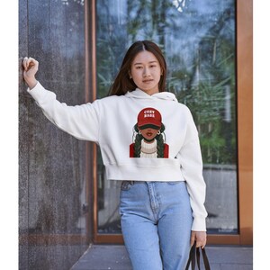 May include: White cropped hoodie with a graphic of a woman wearing a red cap with the text "COZY BABE". The woman in the graphic has braids and is wearing a red jacket. The hoodie is paired with light blue jeans.