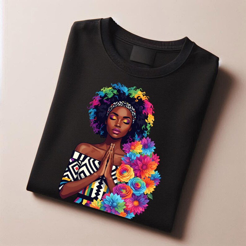 May include: Black t-shirt featuring a vibrant illustration of a woman with colorful hair and flowers. The artwork includes a patterned top and a headband. The woman is depicted with her hands together in a prayer position.