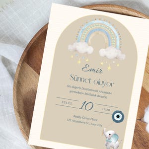 May include: A light blue and yellow rainbow with clouds and stars hangs above the text "Emir Sünnet oluyor". The text below reads "Siz değerli Dostlarımızı Aramızda görmekten Mutluluk duyarız" and "EYLÜL 10 11:30". The text at the bottom reads "Really Great Place 123 Anywhere St., Any City". A blue and white evil eye charm is attached to a string held by a white elephant with blue accents.