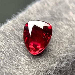 May include: A faceted, heart-shaped, deep red gemstone. The gem has a high luster and reflects light, showcasing its internal structure. The stone is set against a soft, gray, textured background, highlighting its color and shape.