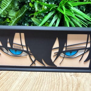 Puede incluir: Una caja rectangular negra con un rostro estilizado de anime con ojos azules y cabello negro. Los ojos tienen reflejos blancos y están sobre un tono de piel claro. La caja está sobre una superficie de madera.
