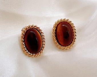 May include: A pair of oval-shaped stud earrings. Each earring features a polished, brown gemstone set within a gold-colored, rope-textured frame. The earrings are displayed on a white, satin-like surface.