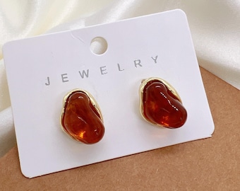 May include: A pair of amber-colored stud earrings with a gold-tone border. The earrings are displayed on a white card with the word "JEWELRY" printed on it. The earrings have a smooth, organic shape.