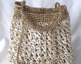 Könnte beinhalten: Eine cremefarbene, gewebte Handtasche mit goldenen Metallic-Akzenten. Die Tasche hat einen Kordelzugverschluss und einen goldenen Kettenriemen. Die Textur der Tasche ist sichtbar, mit einem gewebten Muster und goldenen Verzierungen.