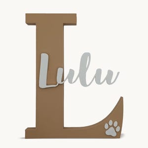 Puede incluir: Una letra "L" de madera de color marrón, con el nombre "Lulu" en fuente cursiva plateada. Una huella de pata blanca está en la esquina inferior derecha. Este artículo decorativo es para dueños de mascotas.