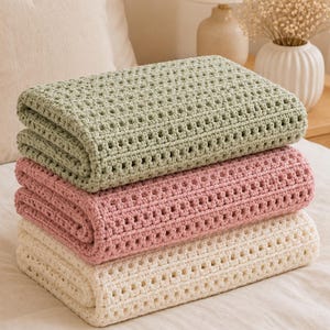 Può includere: Una pila di tre coperte all'uncinetto piegate nei colori verde salvia, rosa antico e crema. Ogni coperta presenta un motivo a griglia strutturato e coerente creato con punti di filato, con una forma rettangolare uniforme e spessa.