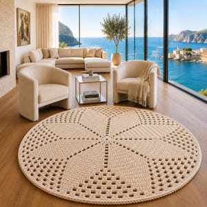 Puede incluir: Una alfombra redonda de ganchillo en color crema con un patrón geométrico repetitivo que irradia desde el centro. El diseño consta de secciones sólidas y caladas alternas, creando un motivo texturizado similar a una flor en toda la superficie.