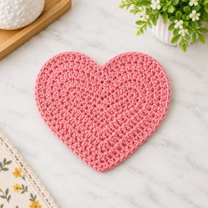 Può includere: Un sottobicchiere a forma di cuore realizzato in filato rosa, caratterizzato da un motivo a uncinetto denso e testurizzato. L'oggetto è piatto e simmetrico, con un colore rosa uniforme su tutta la superficie e lungo i bordi.