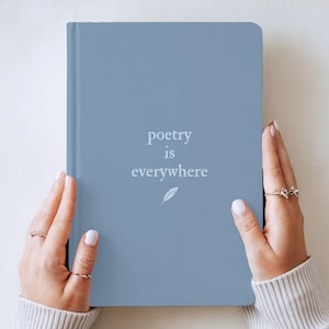 以下が含まれることがあります： 中央に白い文字で「poetry is everywhere」と印刷され、小さな白い羽のアイコンが添えられた、ダスティブルーのハードカバーノート。両手で持たれており、それぞれの薬指には細いゴールドの指輪がはめられている。