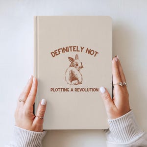 Könnte beinhalten: Ein beiges Notizbuch mit dem Text "DEFINITELY NOT PLOTTING A REVOLUTION" und einer braunen Illustration eines Kaninchens. Das Notizbuch wird von Händen mit Ringen gehalten.