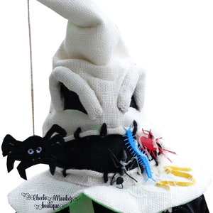 May include: A white felt witch hat with a pointed top and black felt accents. The hat is decorated with black spiders, a blue and red centipede, and a yellow spider web.