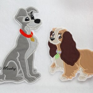 Puede incluir: Dos parches bordados de La Dama y el Vagabundo. El Vagabundo es un perro gris y blanco sentado con un collar rojo. Lady es una perra marr&oacute;n y blanca con un collar morado. Ambos parches tienen un fondo blanco.