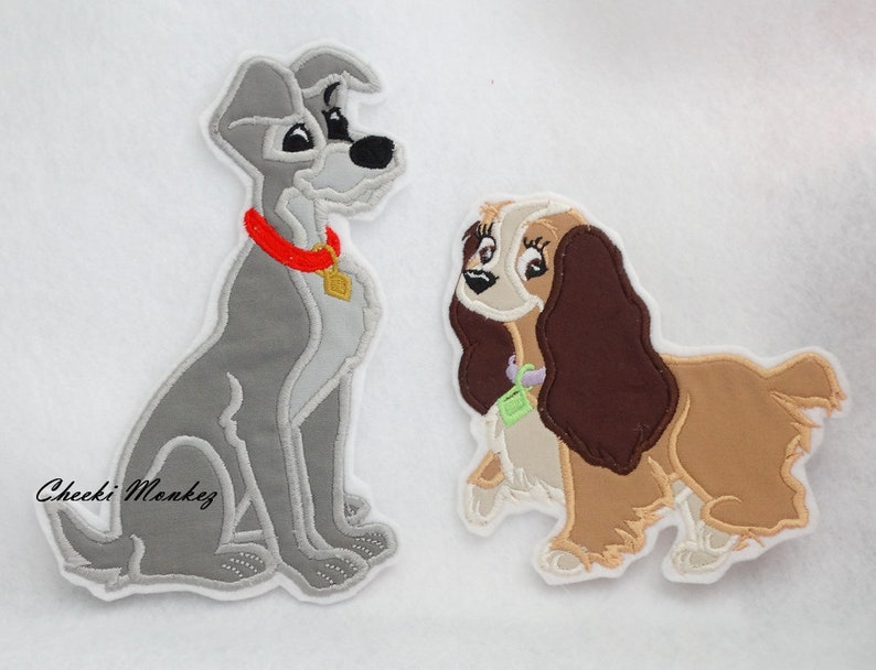 Puede incluir: Dos parches bordados de La Dama y el Vagabundo. El Vagabundo es un perro gris y blanco sentado con un collar rojo. Lady es una perra marr&oacute;n y blanca con un collar morado. Ambos parches tienen un fondo blanco.
