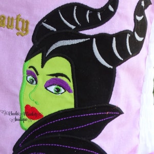 May include: Embroidered patch of Maleficent from Disney's Sleeping Beauty. The patch features a green face with purple eyeshadow and red lips. The character has black hair and horns with silver accents. The patch is on a pink background.