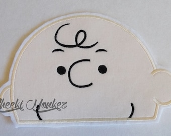 Puede incluir: Aplique de fieltro blanco de la cara de Charlie Brown con rasgos bordados en negro. El aplique tiene forma de semicírculo y tiene un borde blanco.