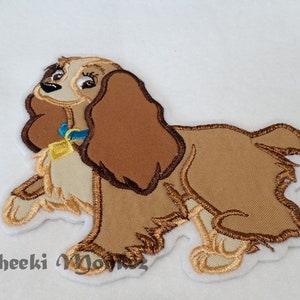 May include: Embroidered patch of Lady, the dog from Disney's Lady and the Tramp. She is brown and white with a blue collar and a gold tag.