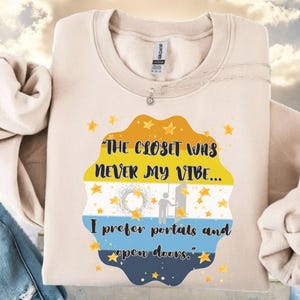 May include: Cream-colored sweatshirt with the text "THE CLOSET WAS NEVER MY VIBE... I prefer portals and open doors." The graphic is a layered design with yellow, blue, and white colors, and star accents.