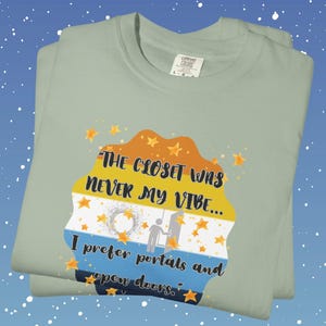 May include: A folded, light green t-shirt with the text "THE CLOSET WAS NEVER MY VIBE... I prefer portals and open doors." The graphic is a colorful design with stars and a silhouette of two people.