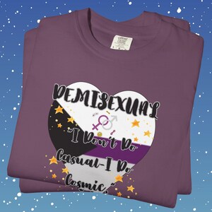 May include: A folded purple t-shirt with a heart-shaped graphic. The graphic features the word "DEMISEXUAL" above the phrase "I Don't Do Casual - I Do Cosmic." The heart design incorporates the demisexual pride flag colors and symbols.