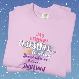 May include: A folded light pink t-shirt with the text "My Gender? Celestial... Floating Somewhere Between Starlight" in various colors. The design includes a crescent moon and stars. The shirt has a Comfort Colors tag.