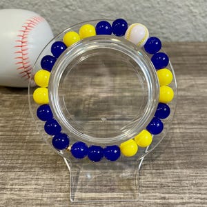 May include: A blue and yellow beaded bracelet with a white baseball charm. The bracelet is on a clear plastic stand.