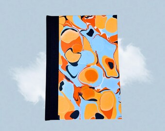 Puede incluir: Un cuaderno de tapa dura con un diseño veteado. La cubierta presenta un patrón de remolinos naranjas, azul claro y negros. El lomo es negro. El cuaderno es rectangular y parece nuevo.