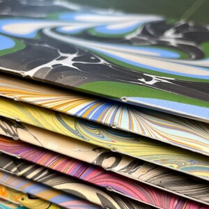 Puede incluir: Una pila de cuadernos con cubiertas de papel jaspeado que presentan patrones de remolinos en varios colores, incluyendo azul, verde, negro, amarillo, rosa y naranja. Cada cuaderno est&aacute; encuadernado con costura de hilo blanco visible en el lomo.