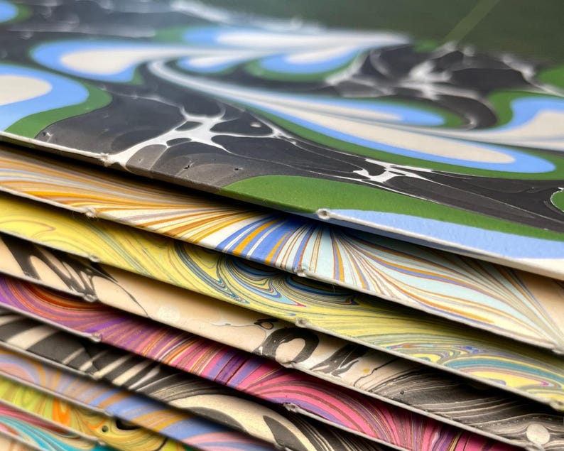 Puede incluir: Una pila de cuadernos con cubiertas de papel jaspeado que presentan patrones de remolinos en varios colores, incluyendo azul, verde, negro, amarillo, rosa y naranja. Cada cuaderno est&aacute; encuadernado con costura de hilo blanco visible en el lomo.