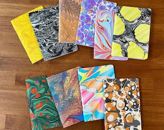 May include: A collection of ten rectangular notebooks featuring diverse marbled paper covers. The designs include patterns in yellow, black and white, tan, purple and blue, red and light blue, yellow and black, green and red, multi-colored, and orange.