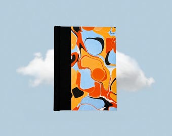 May include: A hardcover notebook with a black spine and a colorful abstract design on the cover. The design features orange, blue, and black shapes against a white background. The notebook is set against a light blue sky with a cloud.