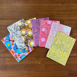 Puede incluir: Una colecci&oacute;n de seis cuadernos rectangulares con cubiertas de papel jaspeado. Los dise&ntilde;os incluyen patrones de remolinos blancos y negros, c&iacute;rculos naranjas y amarillos, ondas moradas y rosas, puntos rosas, remolinos en tonos pastel y ondas amarillas y azules.
