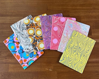 May include: A collection of six rectangular notebooks featuring various marbled paper covers. The designs include patterns of black and white swirls, orange and yellow circles, purple and pink waves, pink dots, pastel multi-colored swirls, and yellow and blue waves.