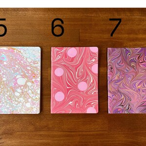 Puede incluir: Tres cuadernos rectangulares etiquetados con los n&uacute;meros 5, 6 y 7. El primero tiene un patr&oacute;n jaspeado en rosa claro, blanco y azul. El segundo es rosa con remolinos blancos y lunares rosas. El tercero presenta un dise&ntilde;o jaspeado morado, rosa y dorado.