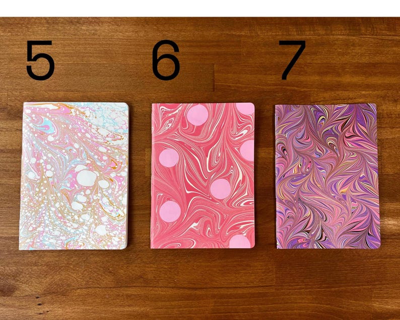 Puede incluir: Tres cuadernos rectangulares etiquetados con los n&uacute;meros 5, 6 y 7. El primero tiene un patr&oacute;n jaspeado en rosa claro, blanco y azul. El segundo es rosa con remolinos blancos y lunares rosas. El tercero presenta un dise&ntilde;o jaspeado morado, rosa y dorado.