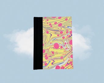 Puede incluir: Un cuaderno con tapa marmolada y lomo negro. La cubierta presenta un fondo amarillo con remolinos rosas y azules. El cuaderno está sobre un cielo azul claro con una nube blanca.