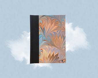 Puede incluir: Un diario de tapa dura con lomo negro y cubierta marmolada. La cubierta presenta un patrón arremolinado en tonos naranja, azul y crema. El diario está sobre un fondo azul claro con formas de nubes blancas.