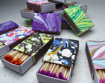 May include: A collection of decorative matchboxes with colorful, marbled patterns. The match heads are various colors, including orange and purple. The boxes are open, revealing the matches inside. The boxes are stacked and scattered on a light-colored surface.