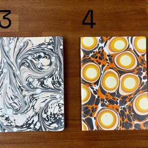 Puede incluir: Dos cuadernos rectangulares etiquetados como 3 y 4. El cuaderno 3 presenta un patr&oacute;n jaspeado en negro, blanco y crema con l&iacute;neas onduladas. El cuaderno 4 muestra un patr&oacute;n vibrante de c&iacute;rculos conc&eacute;ntricos naranjas, amarillos y crema.