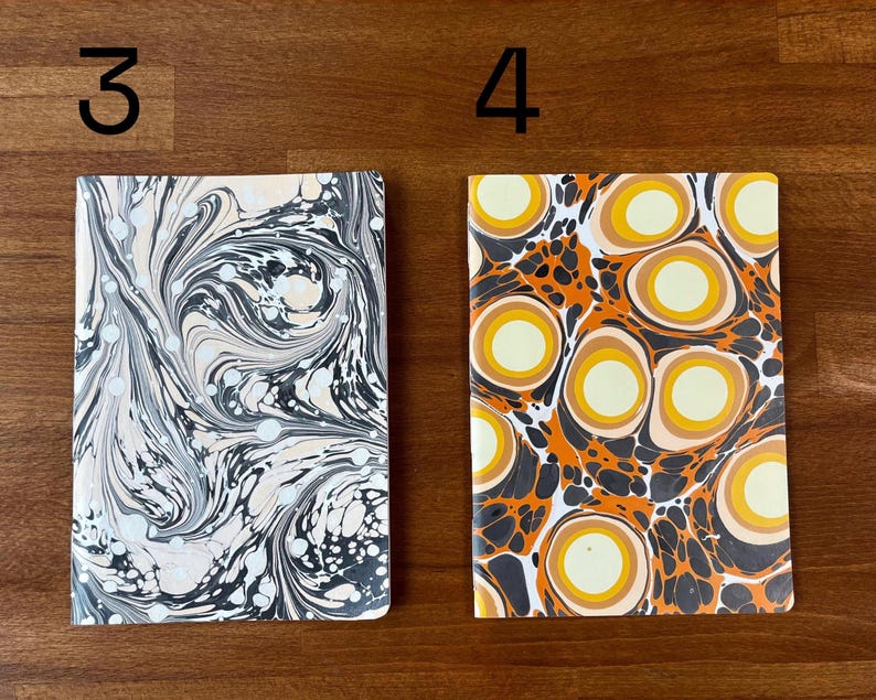 Puede incluir: Dos cuadernos rectangulares etiquetados como 3 y 4. El cuaderno 3 presenta un patr&oacute;n jaspeado en negro, blanco y crema con l&iacute;neas onduladas. El cuaderno 4 muestra un patr&oacute;n vibrante de c&iacute;rculos conc&eacute;ntricos naranjas, amarillos y crema.