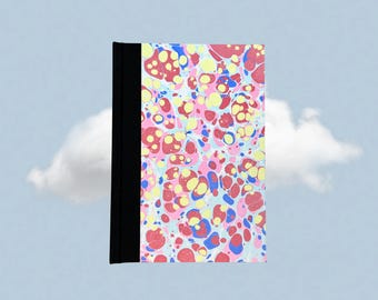 May include: A hardcover notebook with a black spine and a colorful marbled cover. The cover features a pattern of pink, yellow, red, and blue splotches on a white background. The notebook is set against a light blue sky with a white cloud.