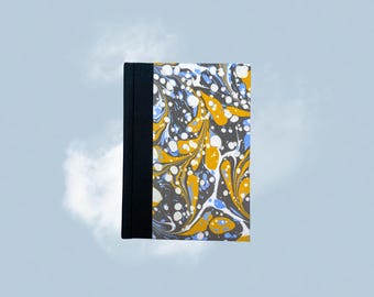 May include: A hardcover notebook with a marbled design in shades of gray, yellow, white, and blue. The spine is black. The notebook is set against a light blue background with white clouds.