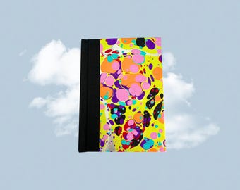 May include: A colorful journal with a black spine. The cover features a vibrant pattern of pink, orange, purple, and black splotches on a yellow background. The journal is set against a backdrop of a blue sky with clouds.