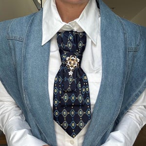 Könnte beinhalten: Ein dunkelblaues Plastron mit einem sich wiederholenden Muster aus gelben und hellblauen Rauten mit zentralen Sternmotiven. Das Plastron wird von einer goldfarbenen Blumenbrosche gehalten, die mit kleinen weißen Perlen und klaren Kristallen verziert ist.