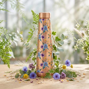 May include: A copper-coloured metal water bottle featuring a repeating diamond grid pattern overlaid with small blue, purple, and yellow floral illustrations. The bottle has a smooth, cylindrical body and a matching metallic copper-toned screw-top lid.