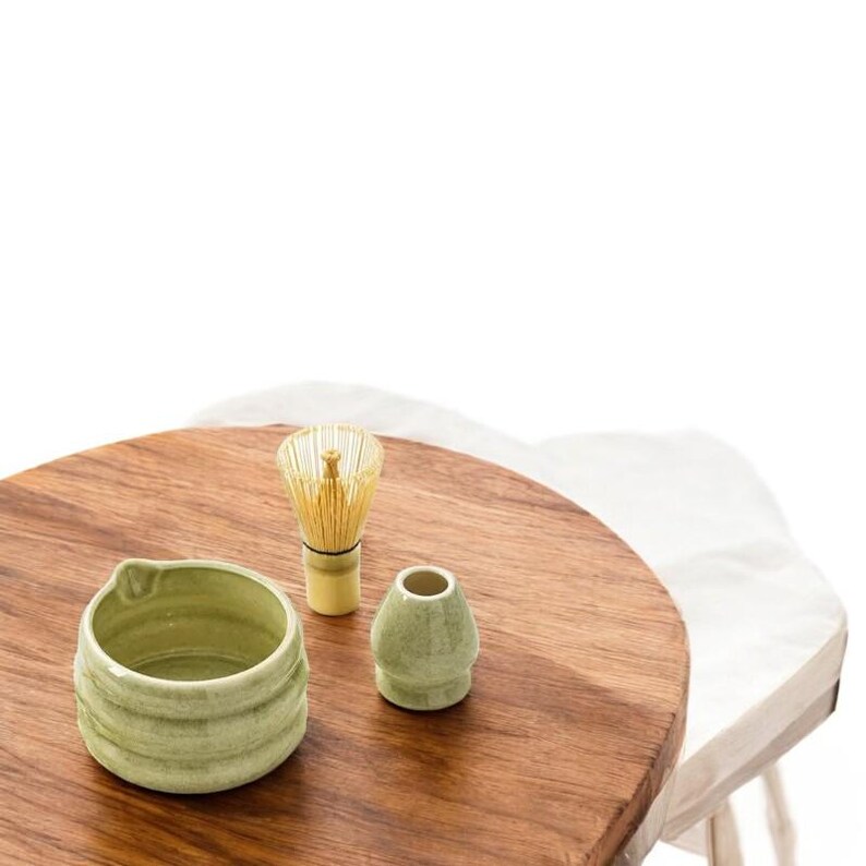 May include: A green ceramic matcha bowl, a green ceramic teacup, and a bamboo matcha whisk on a wooden table.