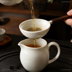 Puede incluir: Una jarra de té de cerámica blanca con asa y pico curvos, que recibe té de color ámbar vertido a través de un colador de té de cerámica blanca con mango de madera oscura. El té fluye en un chorro constante desde el colador hacia la jarra.
