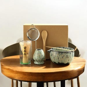 May include: A green and white ceramic matcha bowl with a bamboo whisk, a wooden spoon, a stainless steel strainer, and a box with the text "日本立" in Japanese.