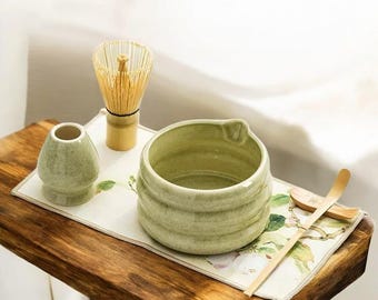 May include: A green ceramic matcha bowl with a bamboo whisk, a small green ceramic cup, and a bamboo matcha scoop on a wooden table with a floral patterned placemat.
