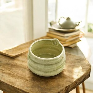 May include: A light green ceramic matcha bowl with a textured, swirling design. The bowl has a slight lip on one side and is sitting on a wooden surface.